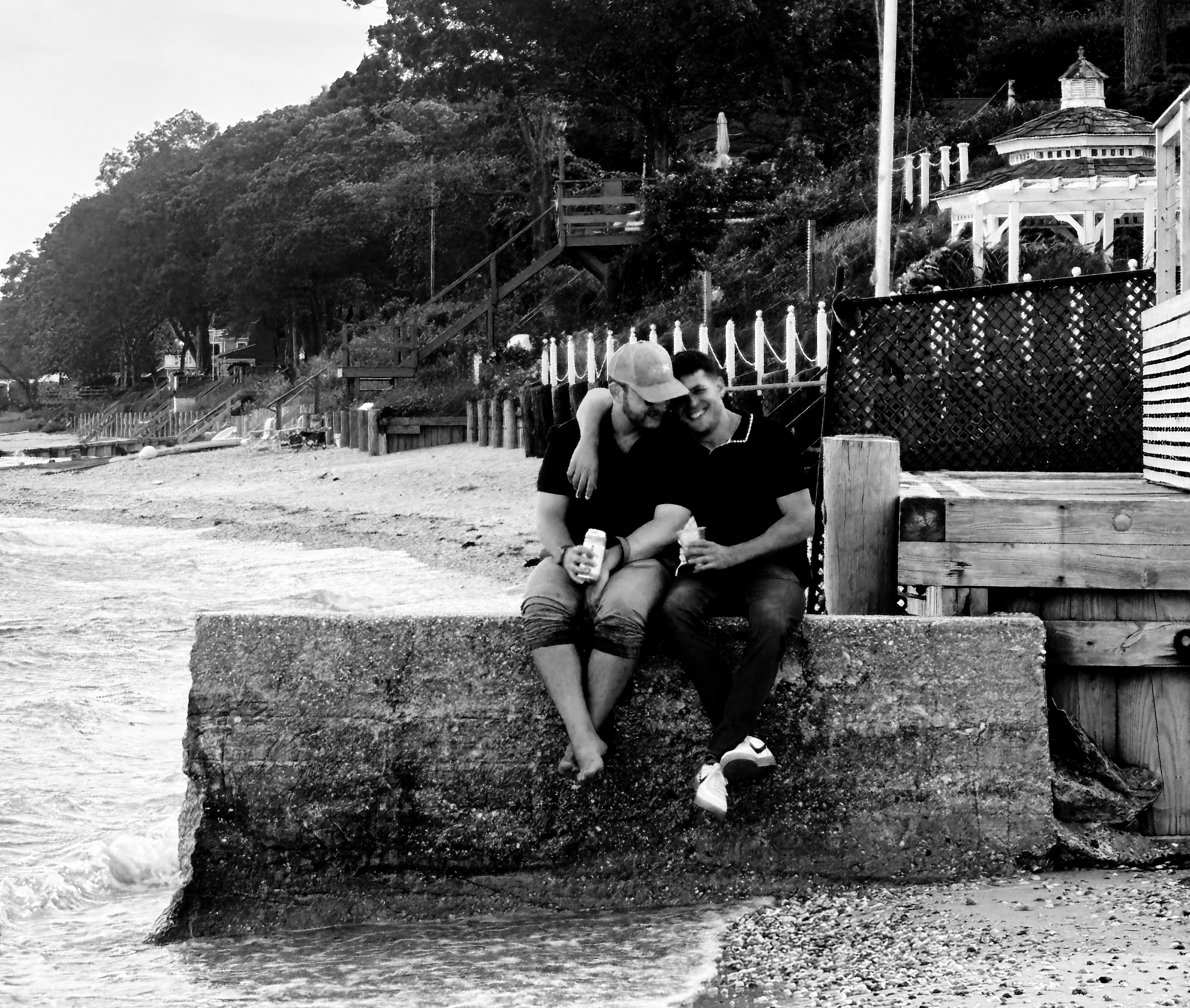 Ian and Zach sitting together on a seawall by the water, black and white
