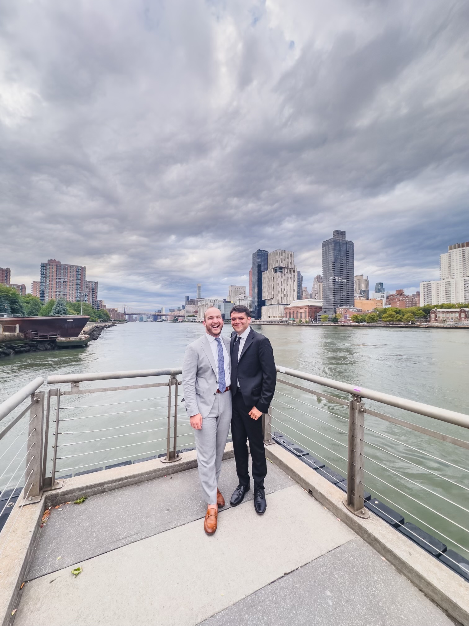 Ian and Zach in suits on a waterfront pier with the New York City skyline behind them